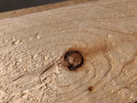 Een kwast omringd door schors, deze mag minder vaak in het hout voorkomen dan een gezonde kwast in de klasses US en V.